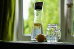 Carafe of water with lemon slices. In front of this are a whole lemon next to a glass mug of water.