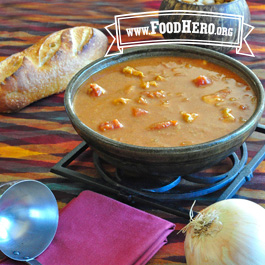 bowl of spicy peanut soup next to a loaf of crusty bread, ladle and fresh onion