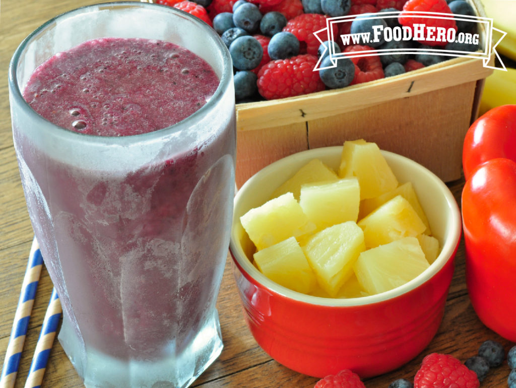 glass of bell pepper smoothie next to small bowl of cut pineapple and box of blueberries and raspberries