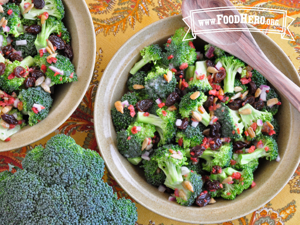 2 bowls of broccoli raisin salad with a fresh broccoli head and wooden spoon