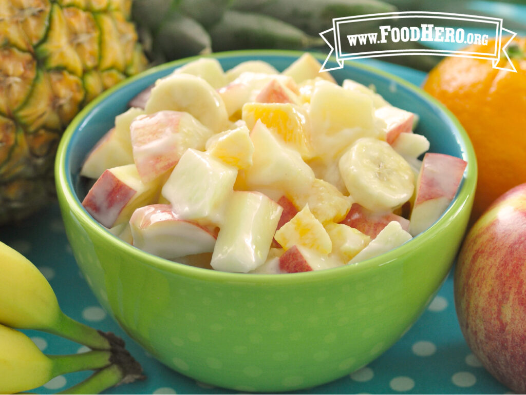 bowl of creamy fruit salad surrounded by apple, orange, pineapple and bananas