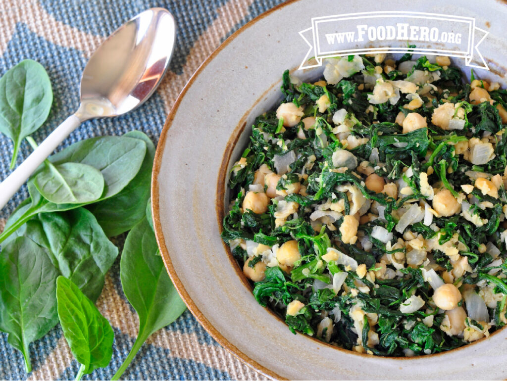 bowl filled with spinach with garbanzo beans next to a spoon and fresh spinach